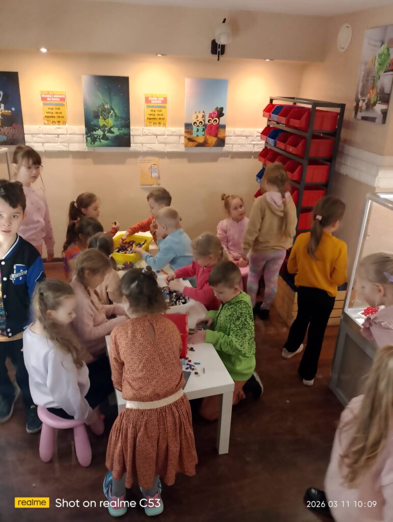 Group of children at white tables building with colorful LEGO bricks in a bright classroom; posters and red bin shelves on the wall behind them.